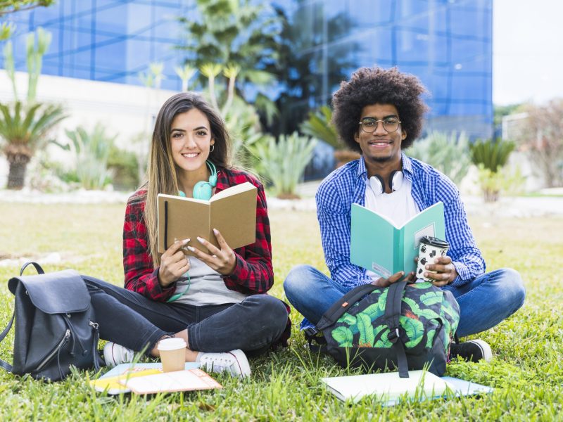diverse-young-couple-holding-books-hand-sitting-lawn-university-campus